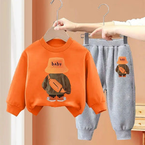 Children's orange sweatshirt and gray pants set with 'baby' design on hangers against a beige background.