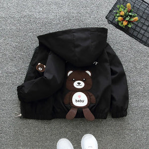 Black hooded jacket with a teddy bear plush toy on a gray floor.