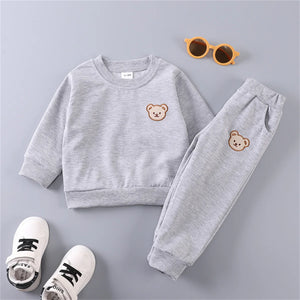 Children's gray sweatshirt and pants set with bear face designs, accompanied by white shoes and orange sunglasses on a light gray background.