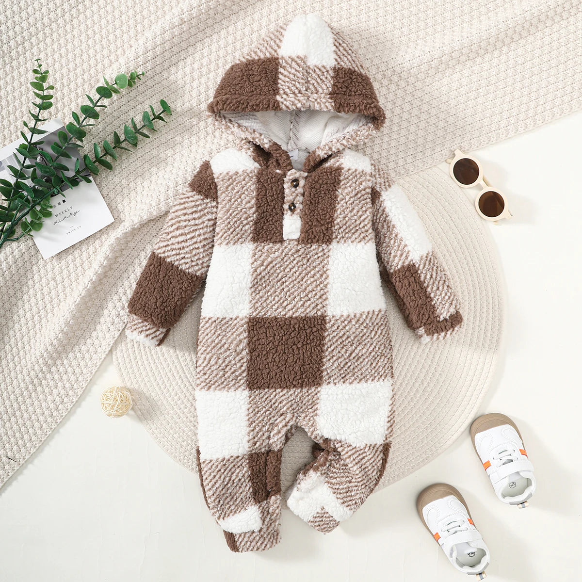 Plaid baby onesie with a hood on a textured surface with shoes and a plant.