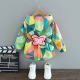 Colorful geometric-patterned jacket with a pink butterfly patch on a hanger against a gray background.