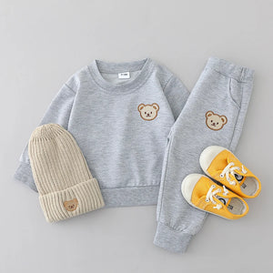 Children's gray sweatshirt, beige beanie, and yellow shoes with teddy bear designs on a light gray background.