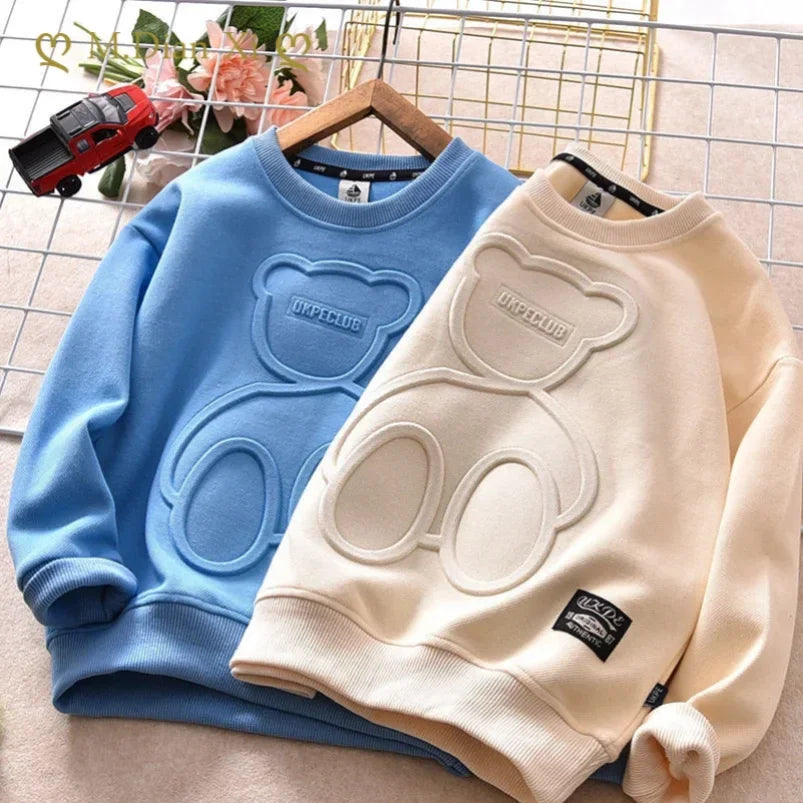 Two sweatshirts, one blue and one beige, with a bear design on a hanger against a grid background.