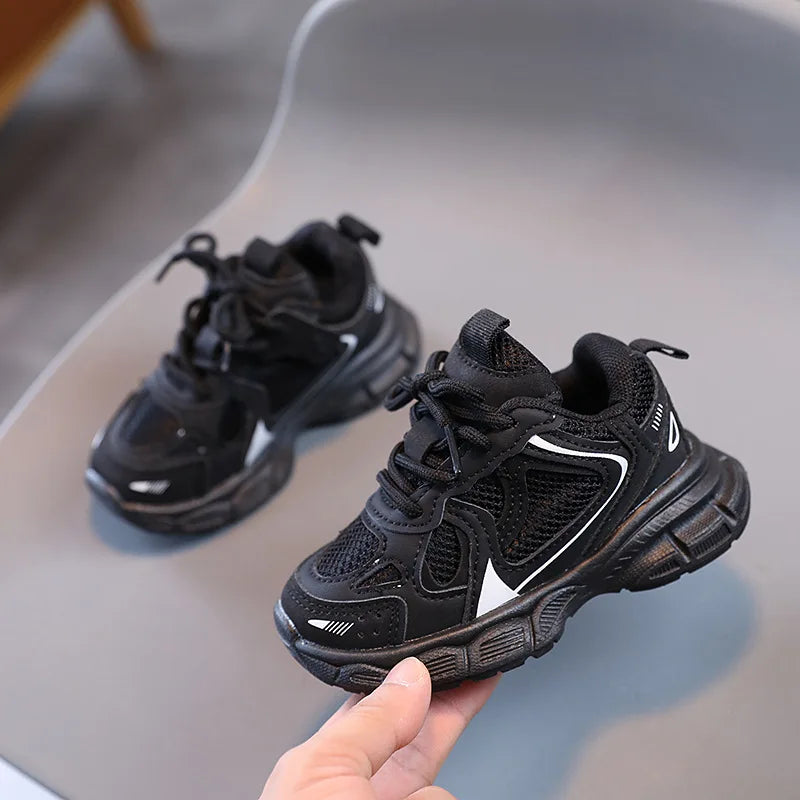 Black children's sneakers on a gray surface with a hand holding one shoe.
