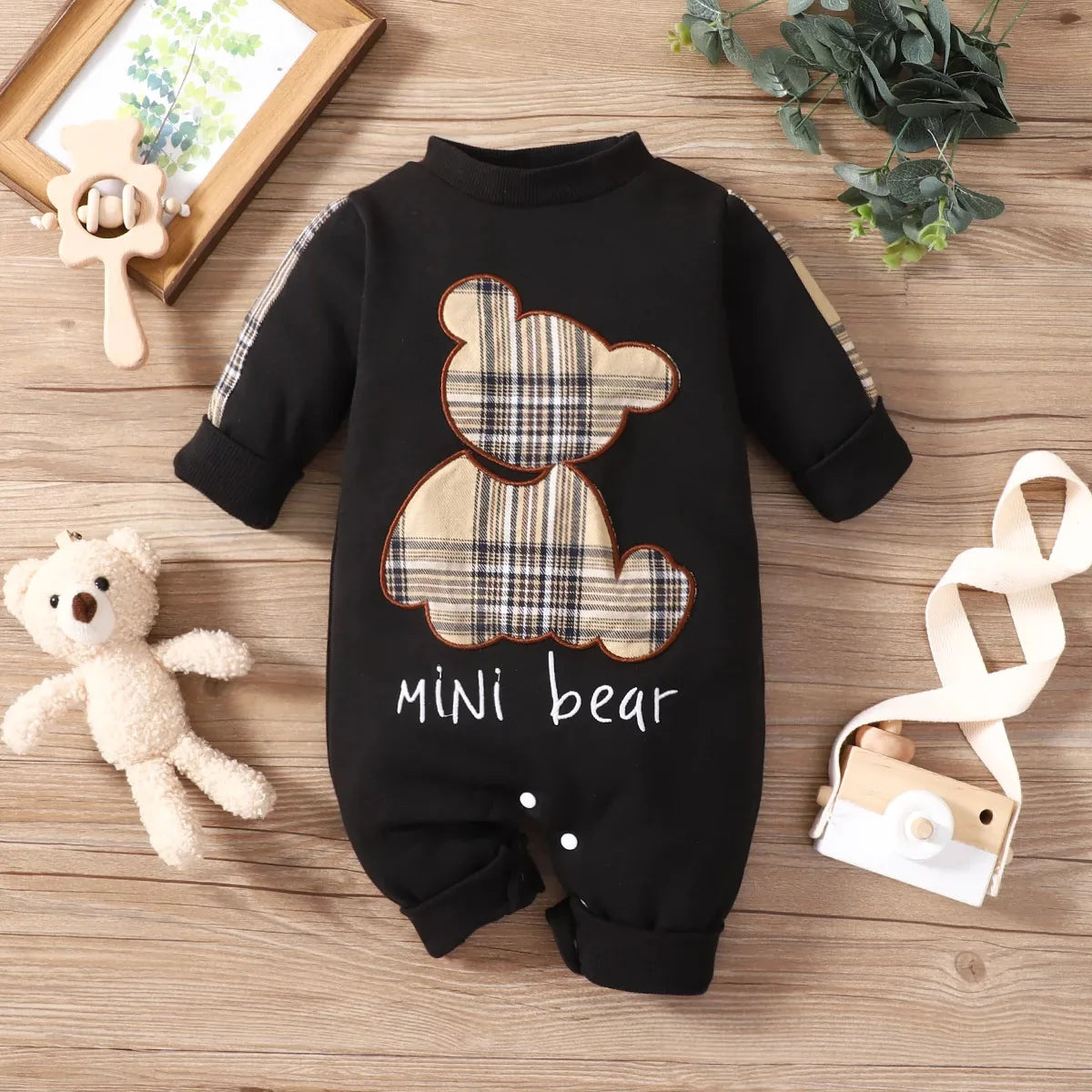 Black baby romper with plaid bear design and 'mini bear' text on a wooden surface with toys.