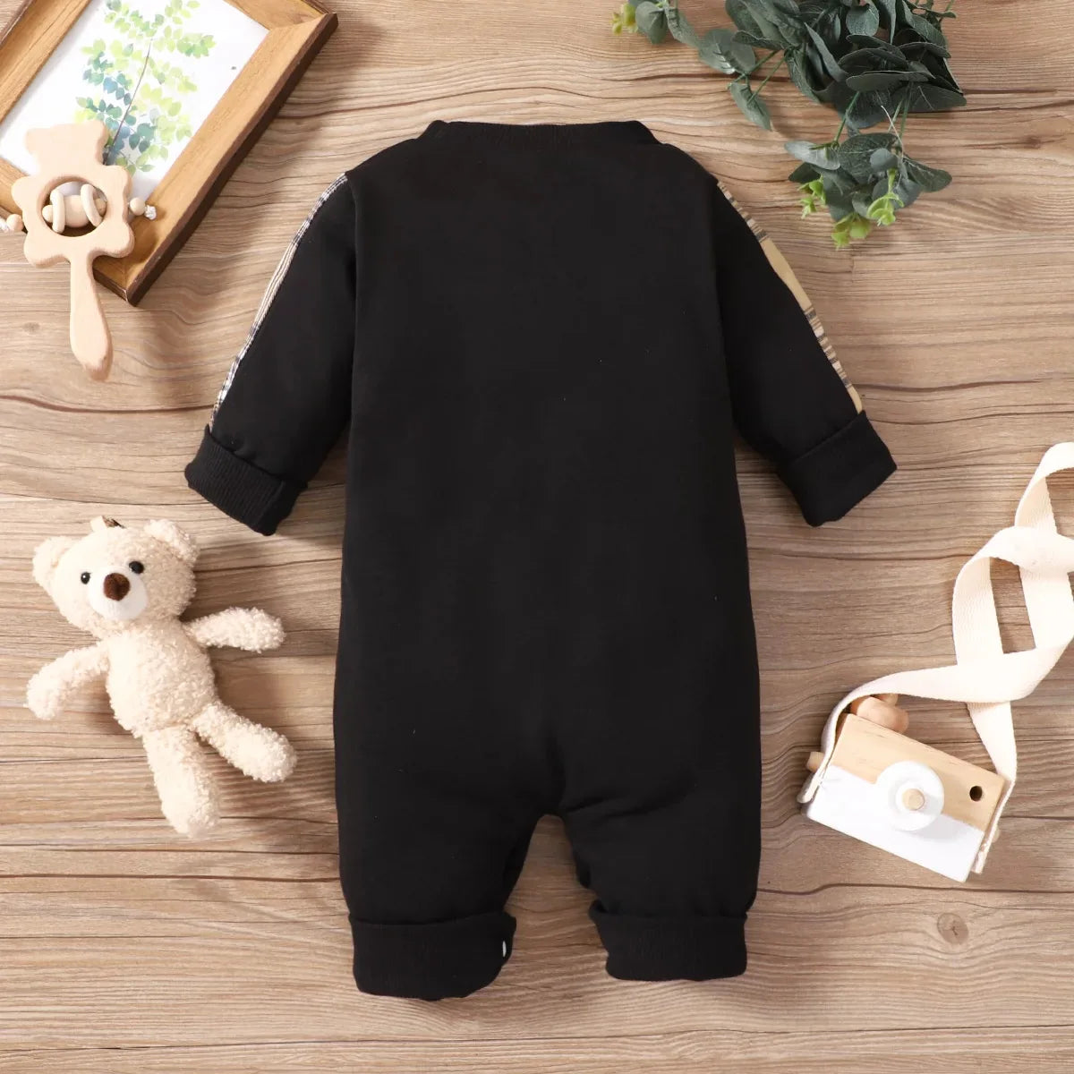 Black baby onesie on a wooden surface with a teddy bear and decorative items.