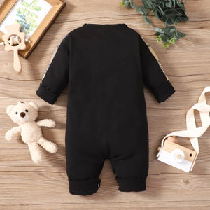 Black baby onesie on a wooden surface with a teddy bear and decorative items.